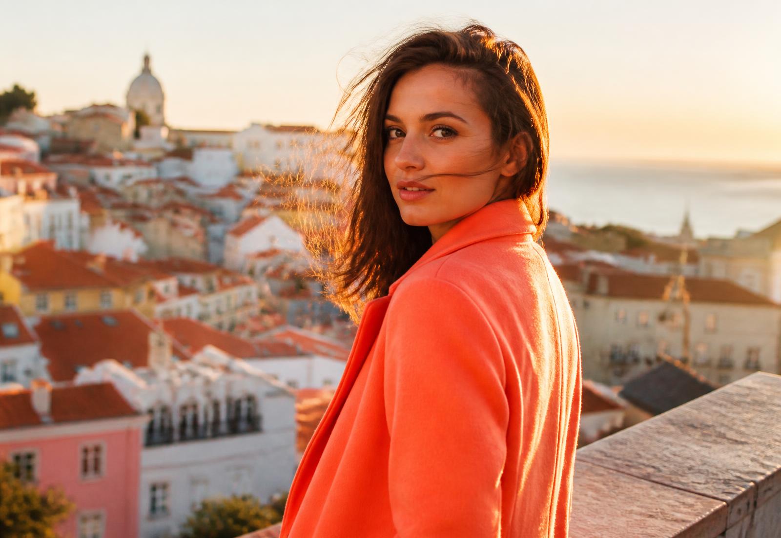 Woman in a bright coral coat on a rooftop above a colourful hillside city at golden hour
