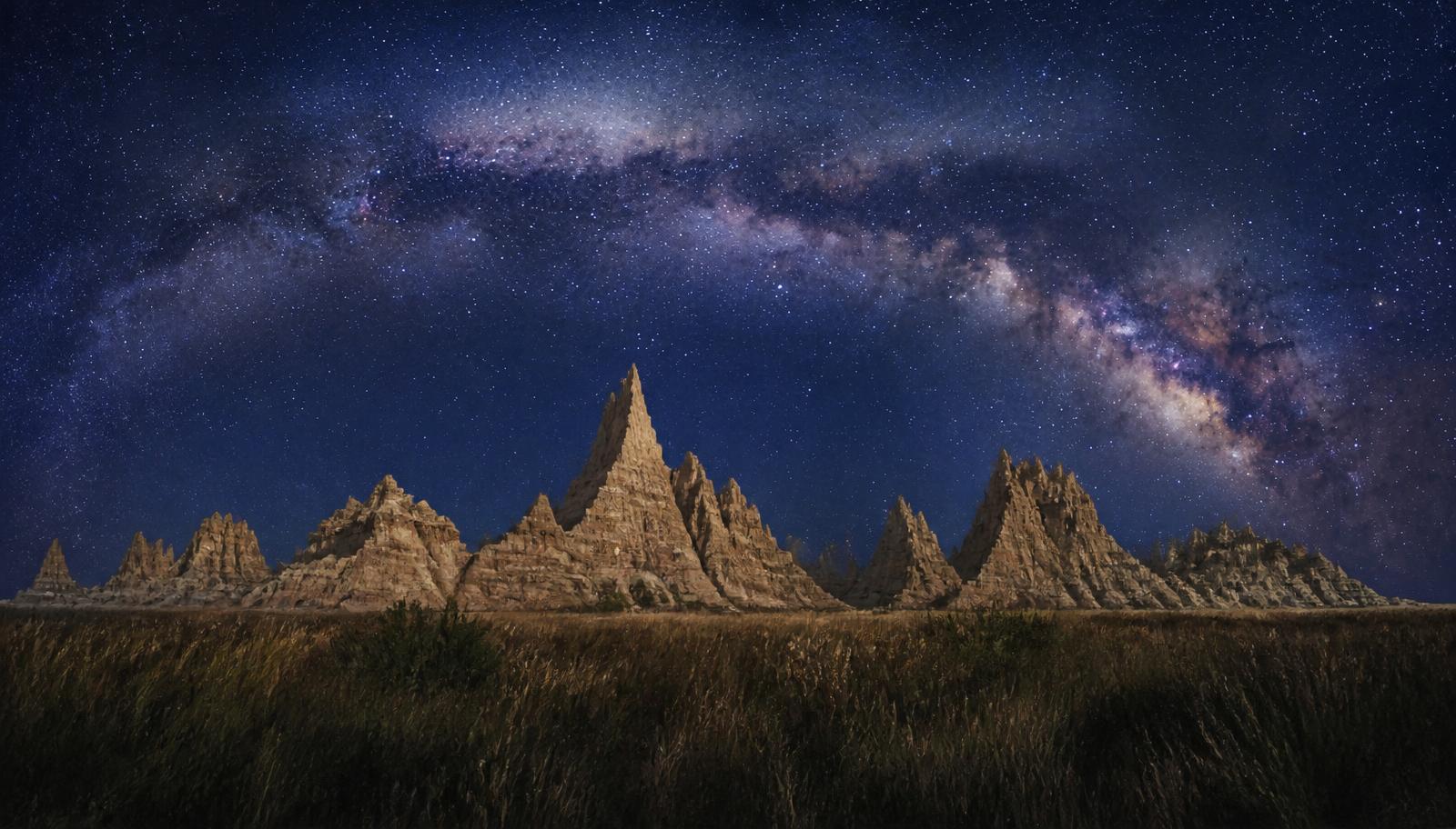 Illustration of the Milky Way arching over eroded badlands rock formations on a clear night